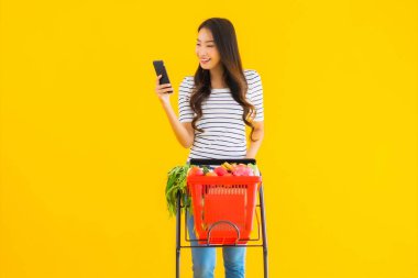Marketten alışveriş yapan güzel Asyalı kadın portresi ve sarı izole edilmiş arka planda akıllı cep telefonu olan market arabası.