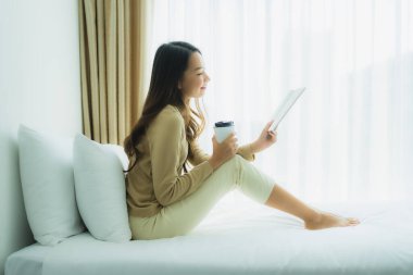 Genç Asyalı kadın, elinde kahve fincanıyla yatak odasında kitap okuyor.