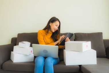 Asyalı genç bir kadının portresi, oturma odasındaki koltukta paket kutusuyla çalışmak için dizüstü bilgisayar kullanıyor.