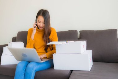 Asyalı genç bir kadının portresi, oturma odasındaki koltukta paket kutusuyla çalışmak için dizüstü bilgisayar kullanıyor.