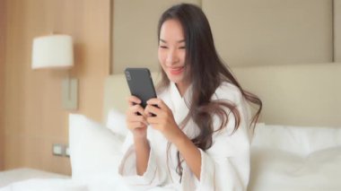 Bornozlu güzel bir Asyalı kadının yatakta otururken akıllı telefon kullandığı görüntüler.