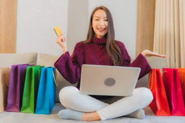 Asyalı güzel bir kadının portresi kanepe üzerinde online alışveriş yapmak için bilgisayarı ve kredi kartını kullanıyor.