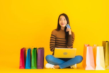 Güzel bir portre genç Asyalı kadın dizüstü bilgisayarla ya da dizüstü bilgisayarla yerde oturuyor sarı arka planda alışveriş torbası etrafında online alışveriş yapmak için kredi kartı kullanıyor.