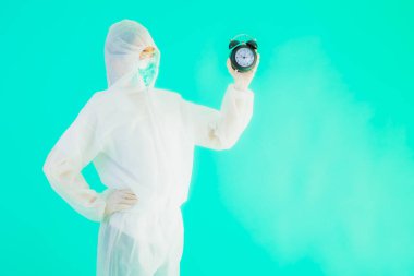Güzel bir portre genç Asyalı doktor. Özel süit ve maske takıyor. Mavi arka planda saati ya da alarmı gösteriyor.