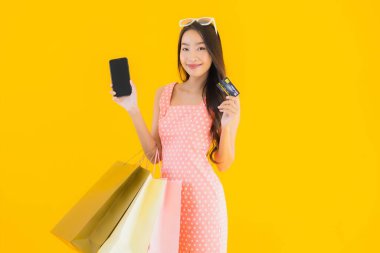 Renkli alışveriş çantalı güzel Asyalı kadın akıllı cep telefonu ve sarı izole edilmiş arka planda kredi kartı.
