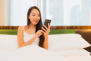 Güzel bir Asyalı kadın yatak odasının içinde akıllı cep telefonu kullanıyor.