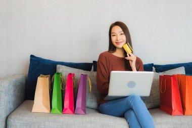Asyalı güzel bir kadının portresi oturma odasındaki kanepede online alışveriş yapmak için bilgisayar ve kredi kartı kullanıyor.