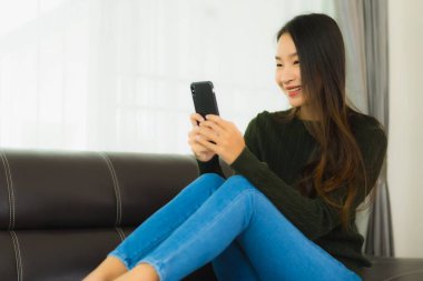 Güzel bir Asyalı kadın oturma odasındaki koltukta akıllı cep telefonu ya da cep telefonu kullanıyor.
