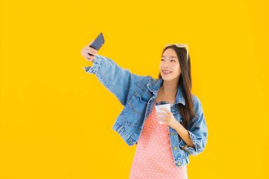 Kahve fincanı ve sarı izole edilmiş arka planda akıllı cep telefonlu güzel Asyalı kadın portresi.