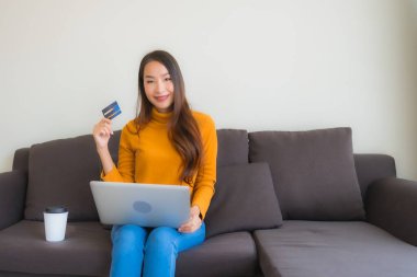 Genç Asyalı kadının oturma odasında online alışveriş yapmak için akıllı cep telefonu ve kredi kartı olan dizüstü bilgisayarı kullandığı portre.