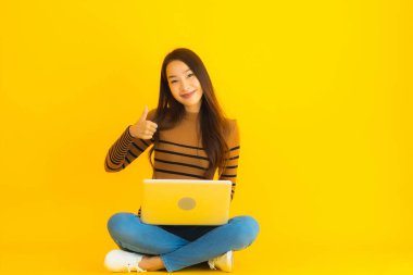 Güzel bir Asyalı kadın portresi, sarı izole edilmiş arka planda dizüstü bilgisayar kullanmak için yere oturur.