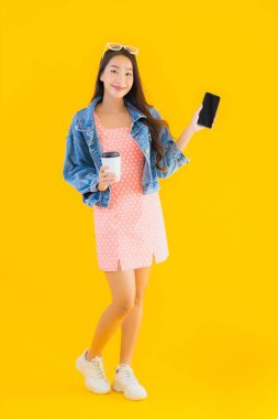 Kahve fincanı ve sarı izole edilmiş arka planda akıllı cep telefonlu güzel Asyalı kadın portresi.