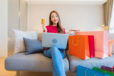 Güzel bir Asyalı kadın portresi. Kanepede çantalı online alışveriş için bilgisayarla kredi kartı kullanıyor.