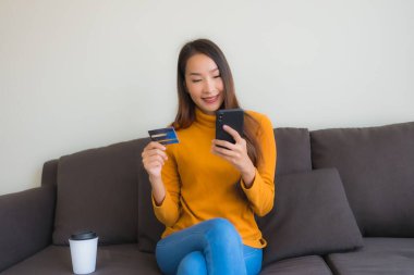 Genç Asyalı kadının oturma odasında online alışveriş yapmak için akıllı cep telefonu ve kredi kartı olan dizüstü bilgisayarı kullandığı portre.