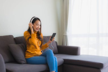 Genç Asyalı kadın oturma odasındaki kanepede müzik dinlemek için akıllı cep telefonu ve kulaklık kullanıyor.