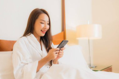 Güzel bir Asyalı kadın yatak odasının içinde akıllı cep telefonu kullanıyor.