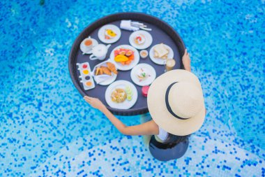 Asyalı güzel bir kadının portresi yüzme havuzunda yüzen kahvaltı tepsisiyle mutlu bir gülümseme