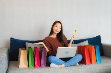 Asyalı güzel bir kadının portresi oturma odasındaki kanepede online alışveriş yapmak için bilgisayar ve kredi kartı kullanıyor.