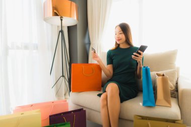 Güzel Asyalı genç kadınların portresi kanepeye oturup bilgisayar bilgisayarı ya da akıllı ve cep telefonuyla online alışveriş için kredi kartı kullanırlar.
