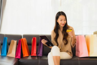 Güzel Asyalı bir kadının portresi. İnternetten alışveriş yapıyor. Kredi kartı ve akıllı telefonuyla oturma odasındaki alışveriş çantasının etrafında.