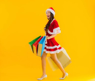 Güzel bir Asyalı kadın portresi. Sarı izole edilmiş arka planda bir sürü alışveriş çantasıyla noel şapkası takıyor.