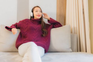 Güzel bir Asyalı kadının portresi oturma odasındaki kanepede müzik dinlemek için kulaklık kullanıyor.