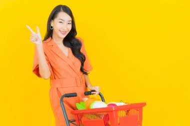 Asyalı güzel bir kadının portresi. Sarı arka planda süpermarketten alınmış alışveriş sepetiyle gülümsüyor.