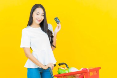 Asyalı güzel bir kadının portresi. Sarı arka planda süpermarketten alınmış alışveriş sepetiyle gülümsüyor.