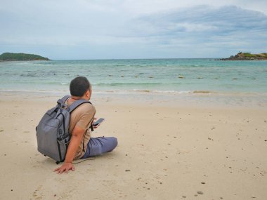 Asyalı şişman backpacker bkz: oturan cep telefonunu güzel gökyüzü ile pastoral plaj tatil zamanında. Samae San Adası Tayland. Yaz konsepti.