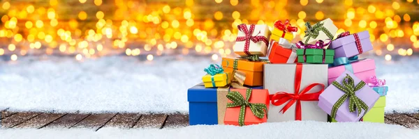 堆五颜六色的圣诞礼物礼物箱子在木板地板雪在明亮的光前面金黄温暖的 Bokeh 宽全景圣诞节背景 — 图库照片