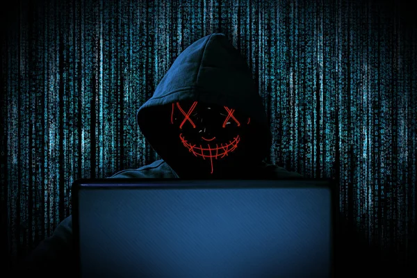Hacker dengan topeng bercahaya merah di belakang laptop notebook di depan — Stok Foto