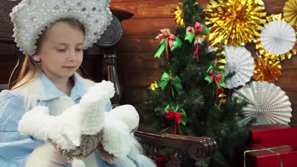 小女孩坐在壁炉附近的木椅上 可爱的好女孩 童话新年的圣诞节角色认为一个软玩具是小狗的小狗 看着我们 闭上你的嘴小雪皇后 — 图库视频影像