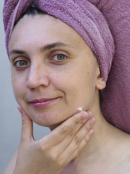 A young woman with a towel on her head smiles and strokes the skin of her face.