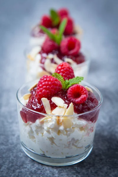 Risalamande is a cold Danish rice pudding served here in three small glass containers in a row on a grey background, as a dessert with cherry jam, flaked almonds, fresh raspberries and sprigs of mint.