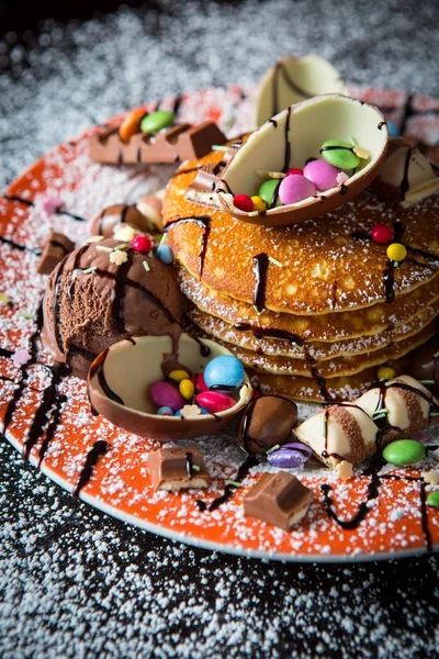 A stack of freshly cooked American butterscotch pancakes drizzled with chocolate syrup and topped with halved chocolate eggs, sweets chocolates on orange rimmed plate on a dark background with sugar