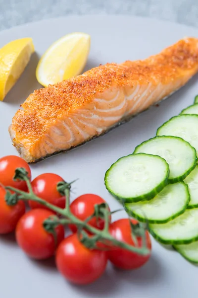 Cooked slice of pink spiced salmon on a white plate with lemon wedges in the background, vine of cherry tomatoes and round slices of cucumber in the foreground.
