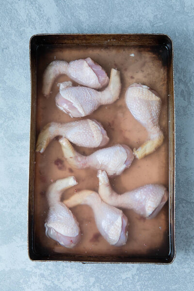 Brined chicken drumsticks in a brining solution in a tray and on a grey background. Top view of brining chicken.