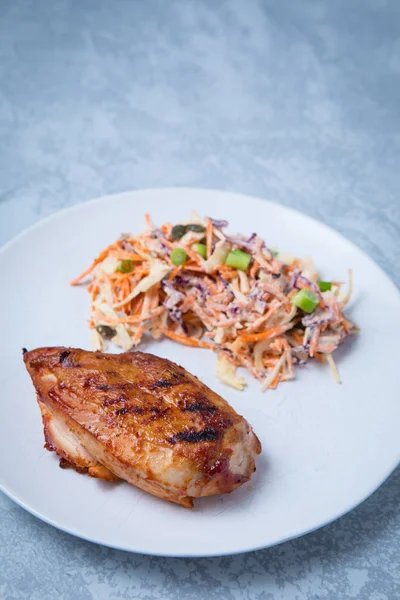 Side view of piece of grilled BBQ chicken breast served with a side grated vegetable salad on a white plate. Grey background.