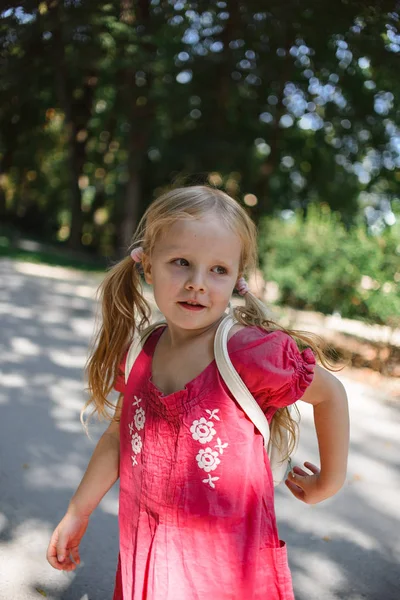cute little blonde girl in red dress with white backpack walking at summer park