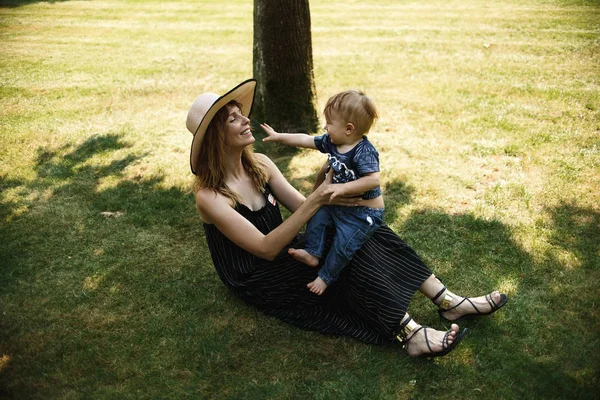 夏の公園で緑の草の上に座っている間 幸せな母親は小さな息子と一緒にプライ — ストック写真