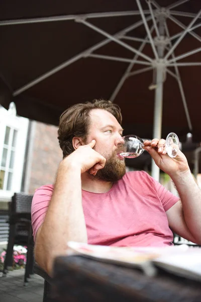 bearded man drinking white wine from glass and sitting in outdoor cafe at summer day