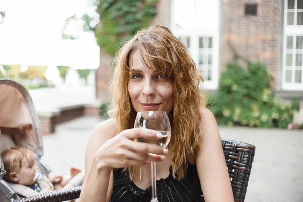 woman with wavy hair drinking white wine from glass and sitting in outdoor cafe at summer day