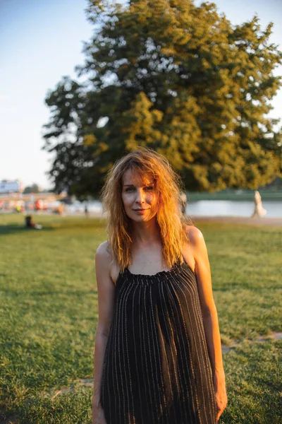 redhead woman with wavy hair in dark dress posing in summer park at sunset