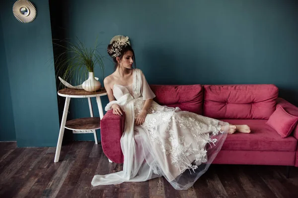 beautiful bride with stylish hairdo in wedding dress sitting on pink sofa