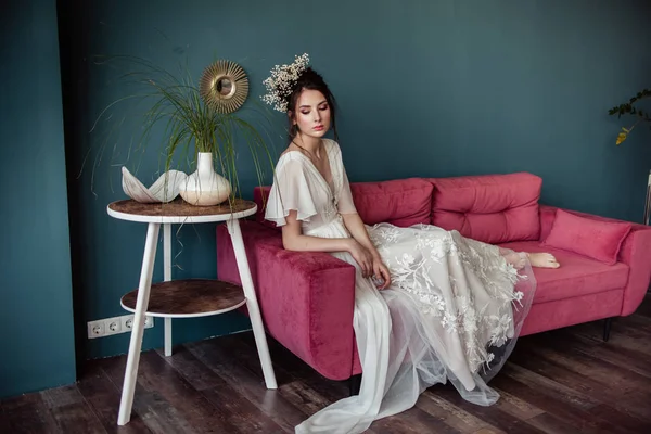 beautiful bride with stylish hairdo in wedding dress sitting on pink sofa