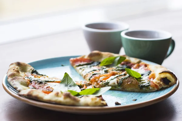 Dilimlenmiş Pizza Margarita veya Margherita bir ahşap masa ve iki fincan çay arka plan karşı mavi tabakta yatıyor. Pizza domates, taze fesleğen, Mozzarella peyniri, biber ve baharat ile kapatın. Geleneksel İtalyan yemekleri.