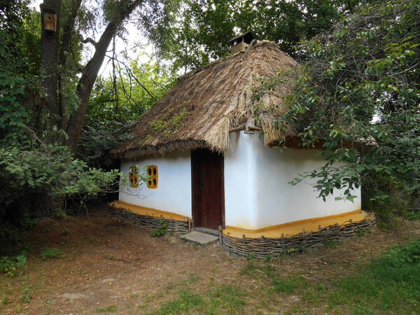 White village house with a thatched roof and a large wooden door, a large courtyard, a cart, a wooden bench, green trees, a blue sky with white clouds