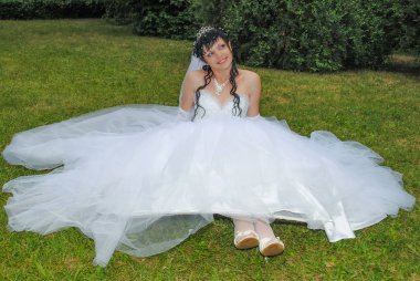 Bir gelinlik gelin çimlerde oturuyor. Beyaz elbiseli bir kız uzakta görünüyor.