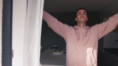 Genç beyaz adam pencereleri açıyor. Yeni günün tadını çıkarıyor. Dinlenmiş ve esniyor.