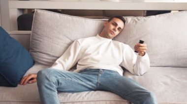 Son derece sıkılmış ve hayal kırıklığına uğramış beyaz bir adam evinde bir kanepede oturup film izlemek için mücadele ediyor, uzaktan kumandayla kanal değiştiriyor.